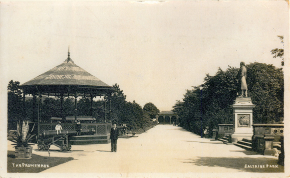 Black and white image showing path through a park with bandstand to left and monument to right of path. Caption reads "The Promenade. Saltaire Park"